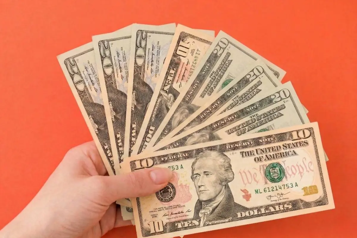 Mano sujetando un abanico de billetes de dólares sobre fondo naranja, representando el pago del Bono de Desarrollo Humano en Ecuador.